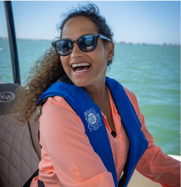 Person smiling on a boat wearing sunglasses, a coral shirt, and a blue life jacket with water in the background.
