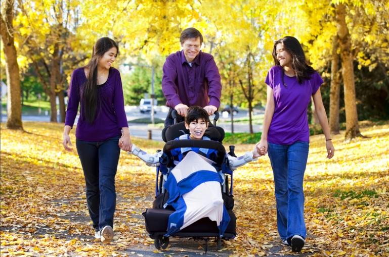 A child in a wheelchair is pushed by a man while holding hands with two women on a leaf-covered path surrounded by yellow autumn trees.