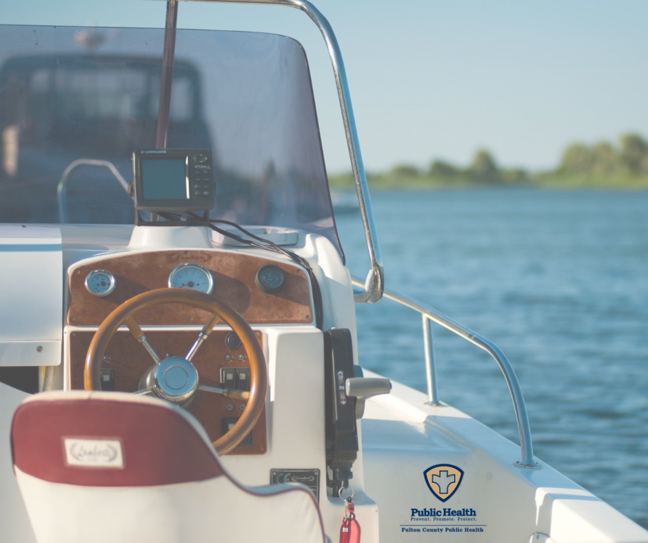 Boat's steering area with a wooden wheel, electronic screen, and maroon seat with water and tree line in the background.