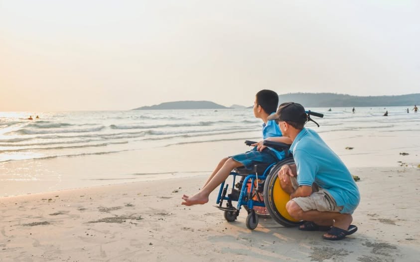 A child in a blue wheelchair watches the ocean at sunset, accompanied by a man in a blue shirt and hat.