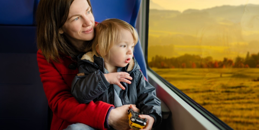 A woman in a red jacket holds a child with blonde hair while traveling on a train, both gazing out a window at a golden landscape.